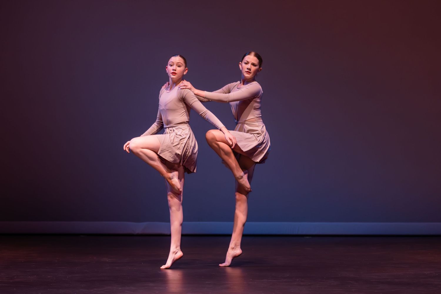 Two dancers in beige costumes perform a synchronized pose with raised legs on a darkly lit stage.