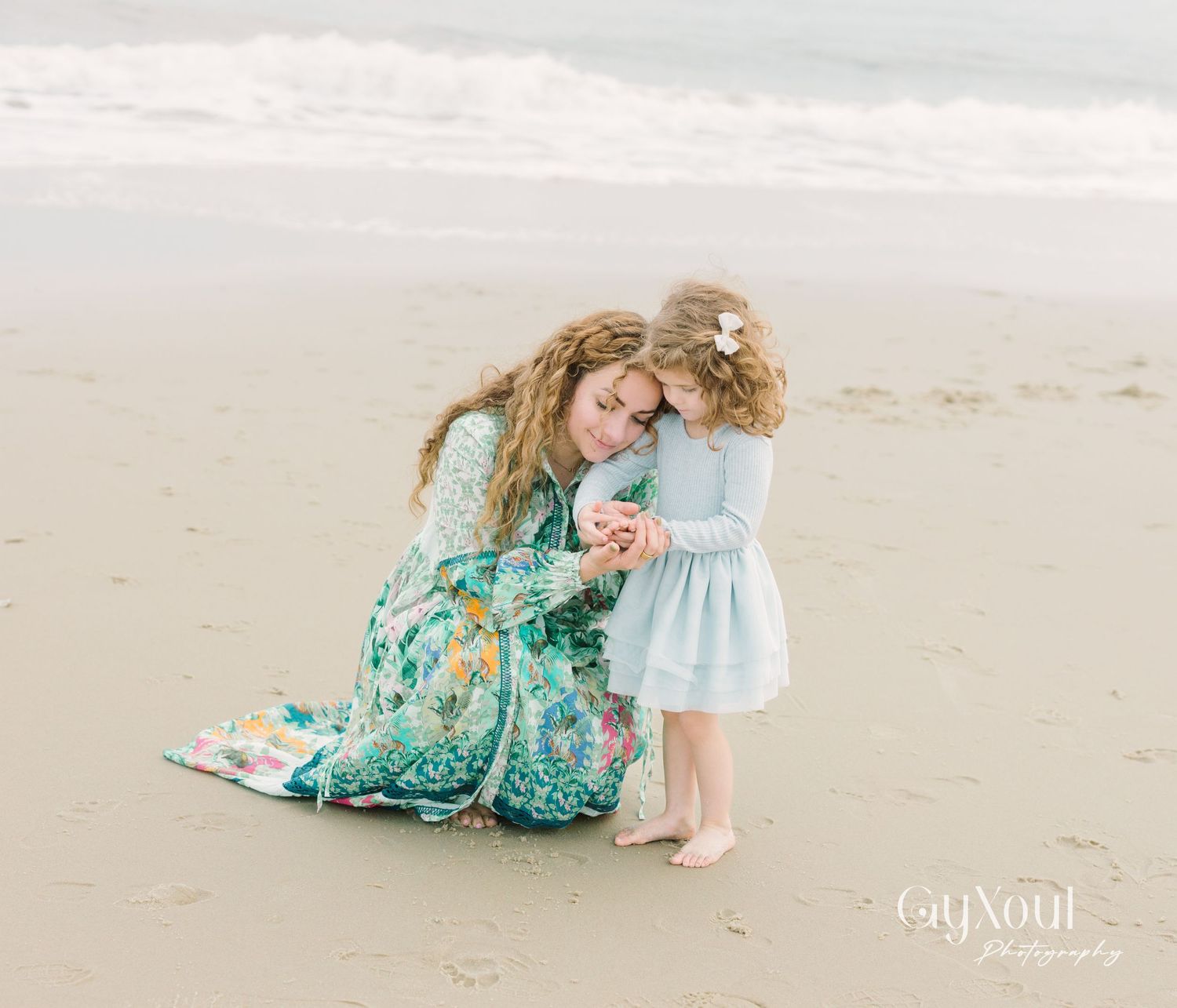 A tender moment shared between two figures wearing matching mint and floral dresses on a sandy beach.