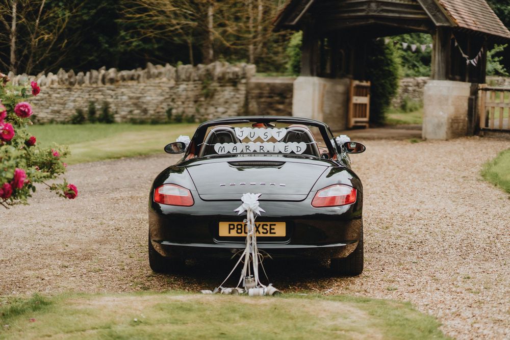 Sleek black Mercedes convertible decorated with wedding ribbons and cans parked on gravel driveway near stone wall and roses.