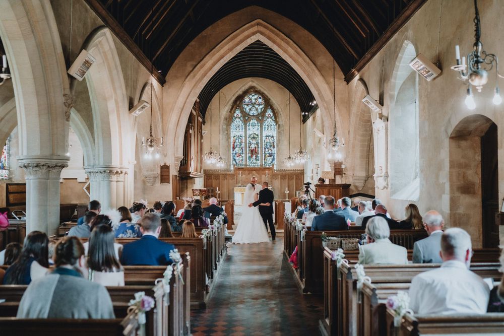 A traditional church wedding ceremony in progress with guests seated in wooden pews beneath gothic arches and stained glass.