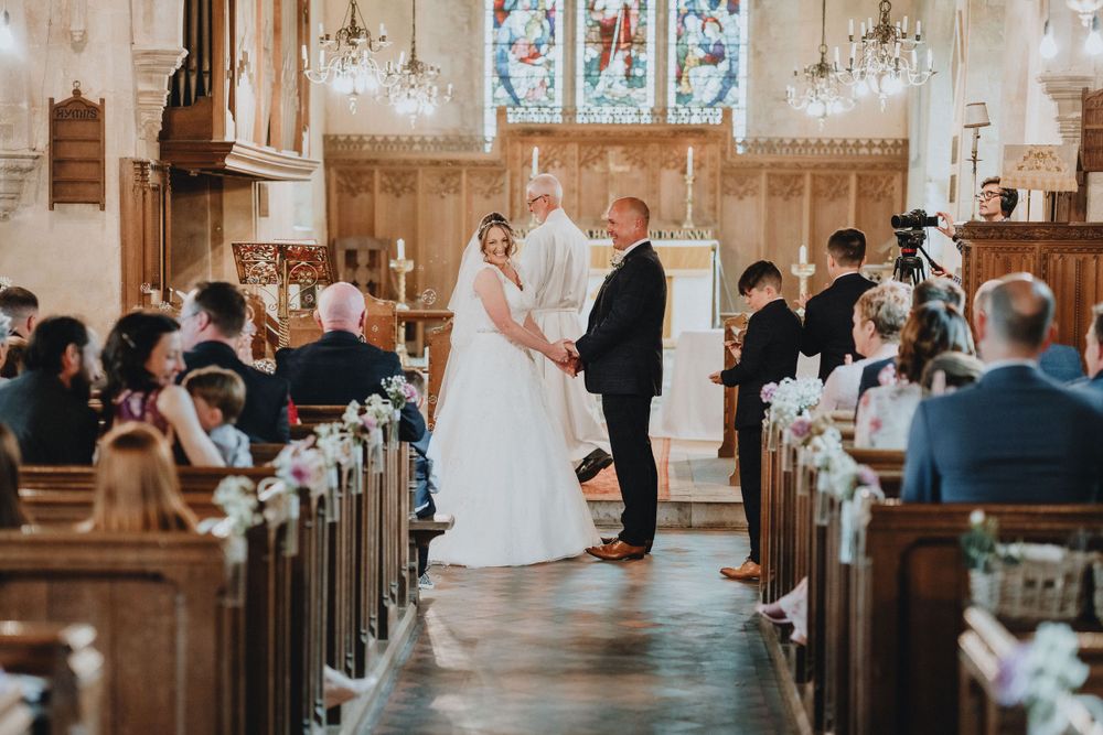A wedding ceremony takes place in a beautiful church with stained glass windows and wooden pews filled with guests.