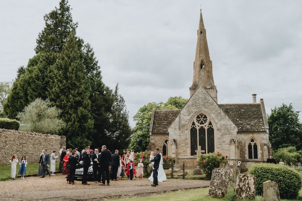 Small stone church with spire hosts outdoor wedding ceremony gathering in countryside setting with overcast sky.