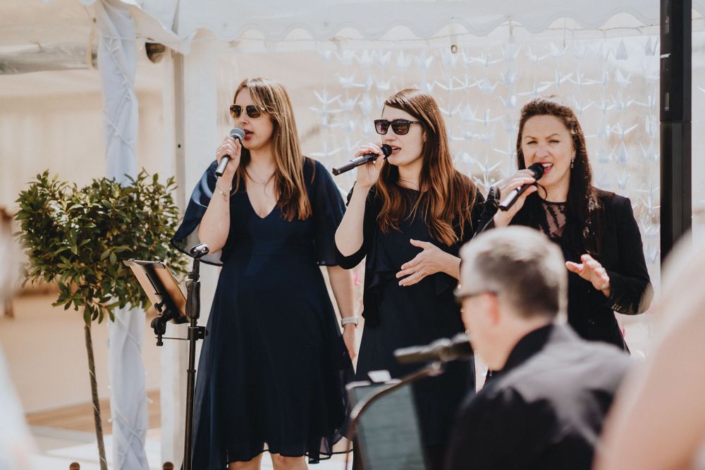 Three performers in black dresses singing together at an event while accompanied by musicians.