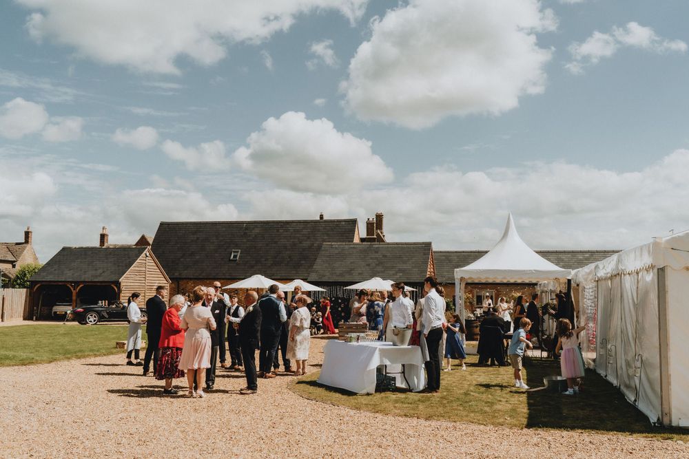 Outdoor market event with white vendor tents and crowd gathered on gravel path near rustic stone buildings under blue sky.