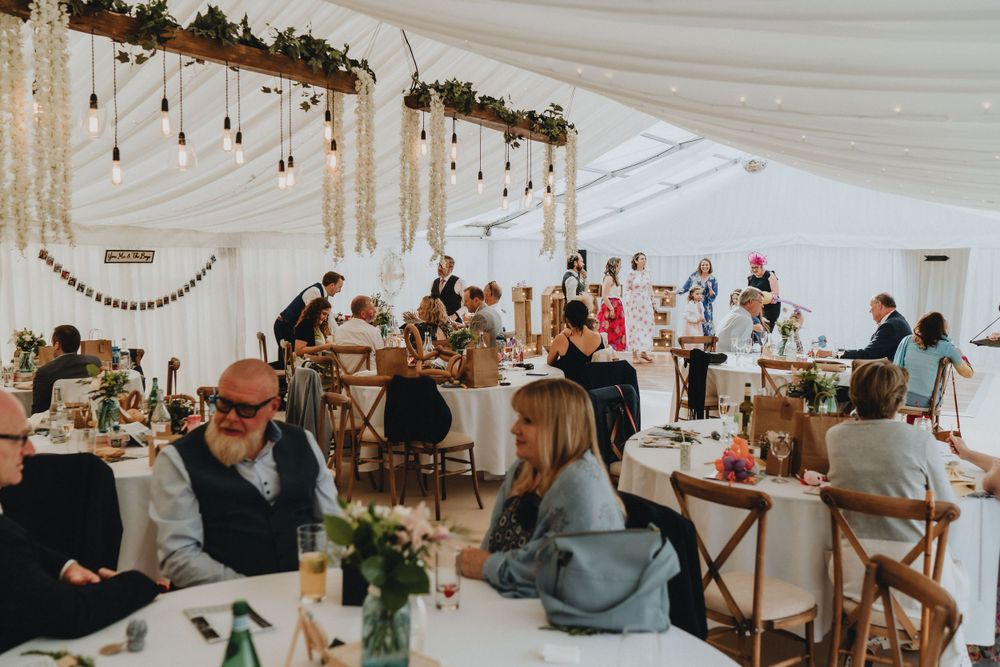 Wedding reception under a white tent with string lights, rustic decor and guests seated at tables with wooden chairs.