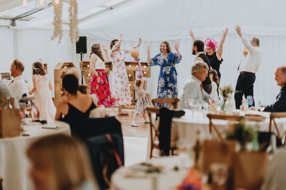 Party guests dancing enthusiastically together under a white tent at an outdoor reception event.
