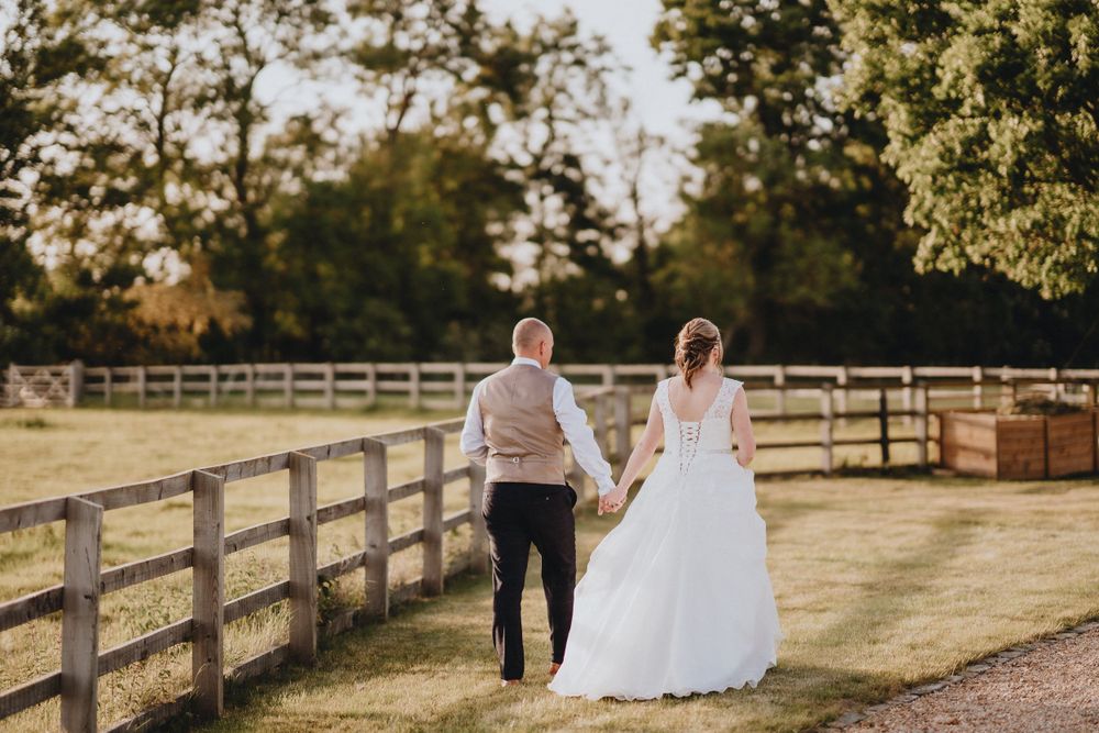 A couple holding hands walks along a wooden fence in a golden sunset at a rustic countryside venue.