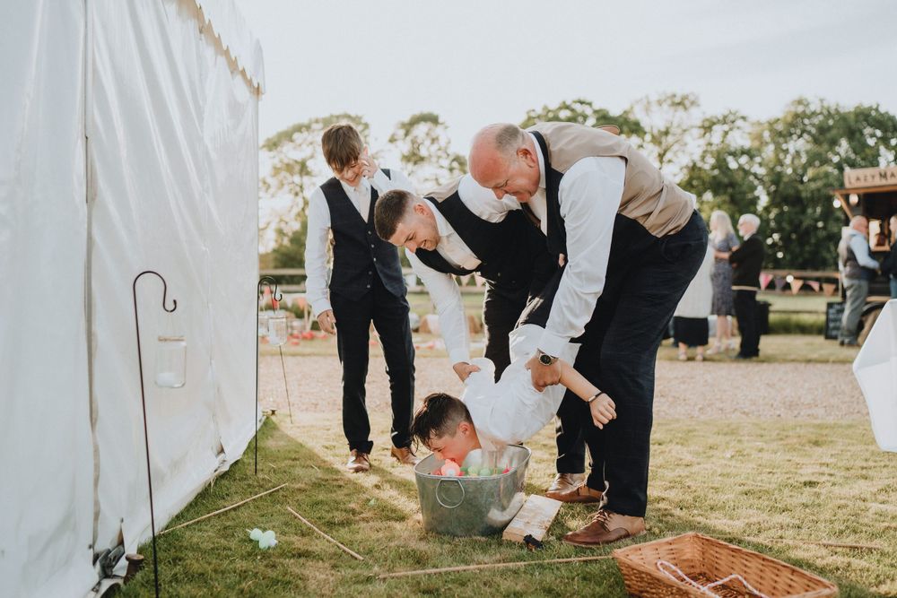 Wedding guests help a small child peeking under a white tent at an outdoor reception.
