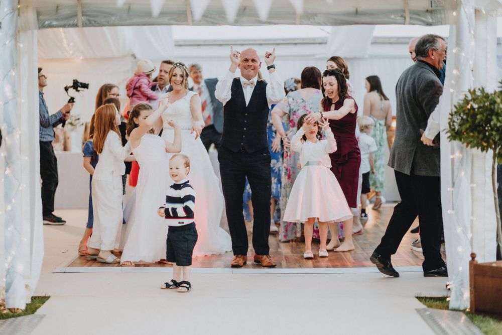 Guests gather under a white-draped outdoor pavilion during a wedding celebration on a bright sunny day.