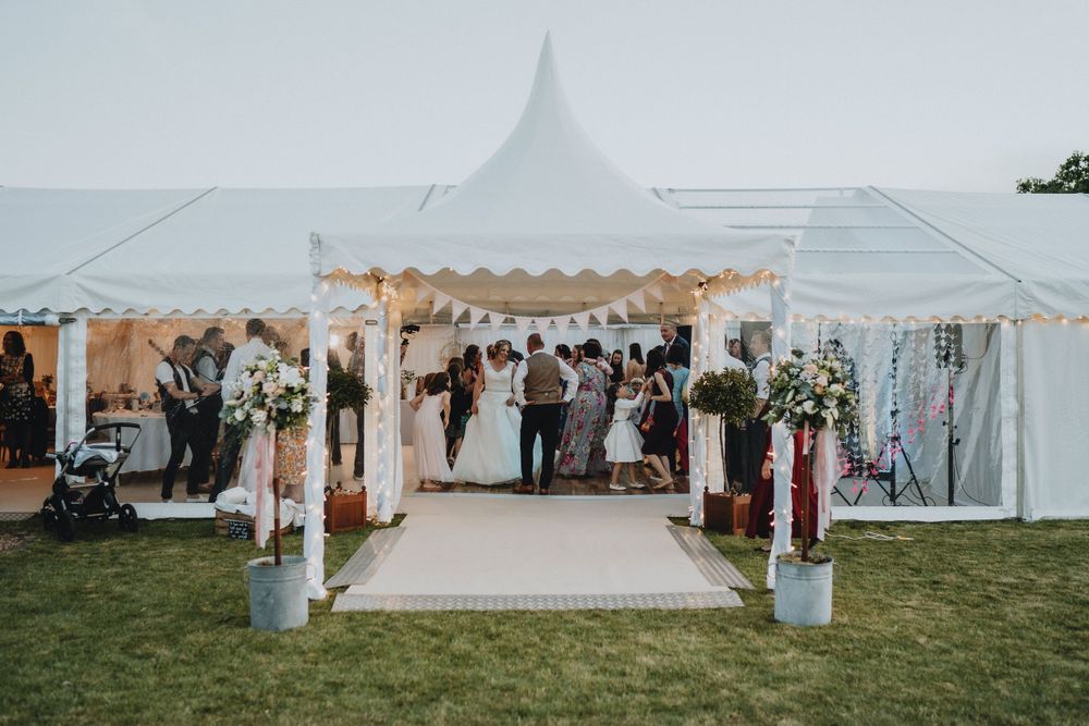 Wedding celebration under an elegant white tent with floral decorations and a raised platform on a grassy lawn.