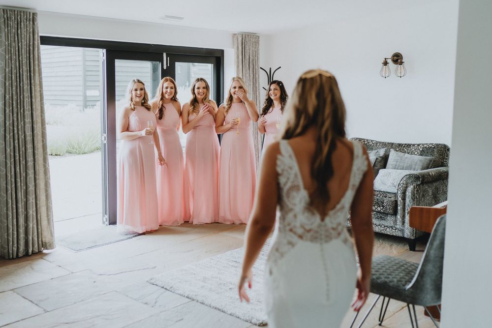 Bridesmaids in pink floor-length dresses react with delight as they look at someone in a white lace wedding gown.
