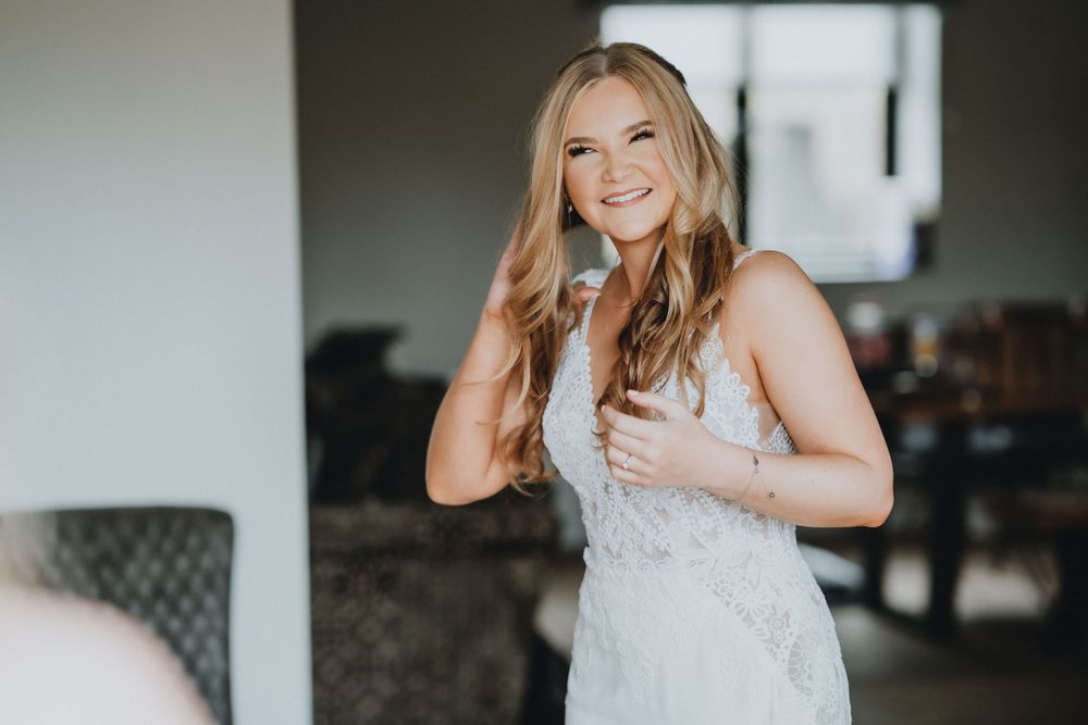 Smiling bride in white lace wedding dress stands indoors while touching her hair and holding a glass.