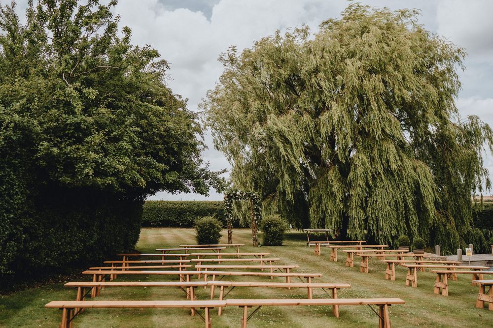 Wooden benches arranged in rows outdoors under a large weeping willow tree for an outdoor ceremony at Lattenbury Hill, Cambridgeshire.
