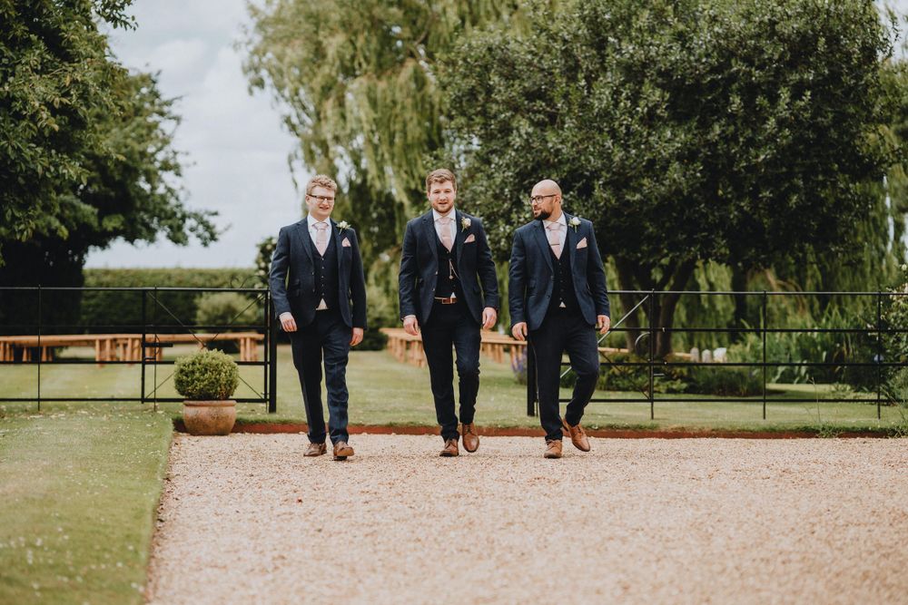Three groomsmen in dark suits walk together on a gravel path in a garden setting.