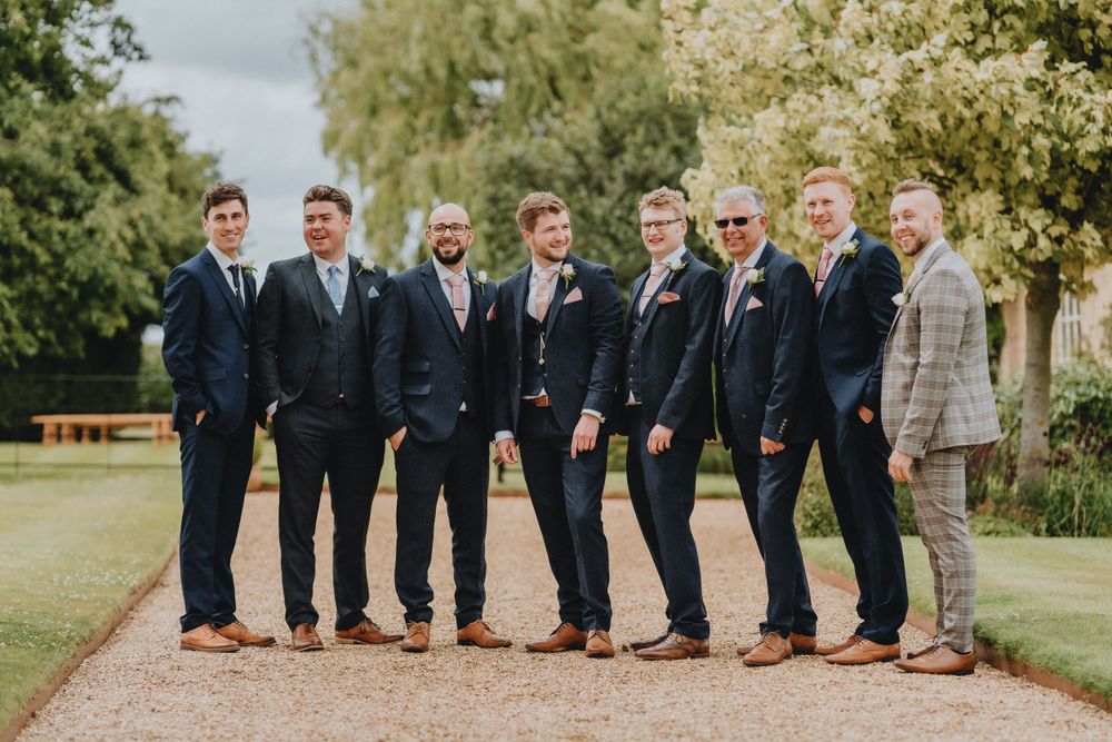 A wedding party in matching dark suits and brown shoes poses together on a gravel path outdoors.