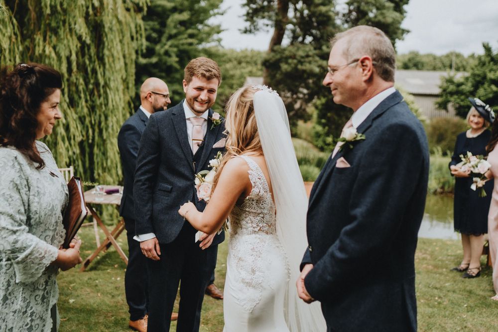 Outdoor wedding ceremony takes place by a willow tree beside a lake on a sunny summer day.