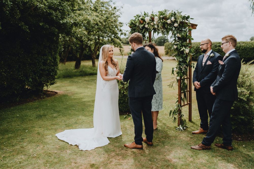 An intimate outdoor wedding ceremony takes place under a wooden arch adorned with greenery on a cloudy summer day.
