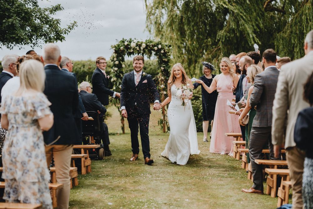 A joyful outdoor wedding ceremony under leafy trees with rustic wooden bench seating and floral arch decorations.