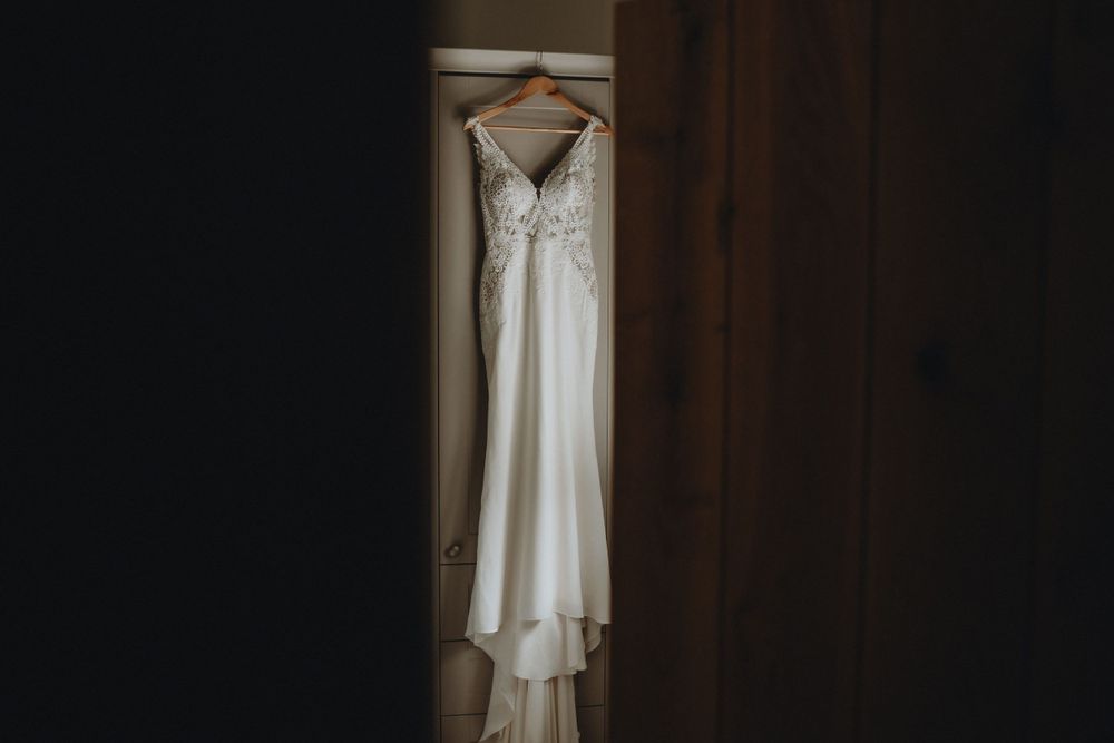 A white wedding dress with delicate lace details hangs elegantly against a dark brown wall in soft lighting.