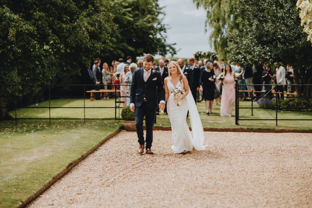 A couple walks down a gravel path at an outdoor wedding ceremony while guests look on from behind.