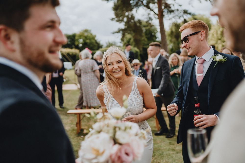 A sunny outdoor wedding celebration with guests gathered on a green lawn, featuring elegant floral bouquets.