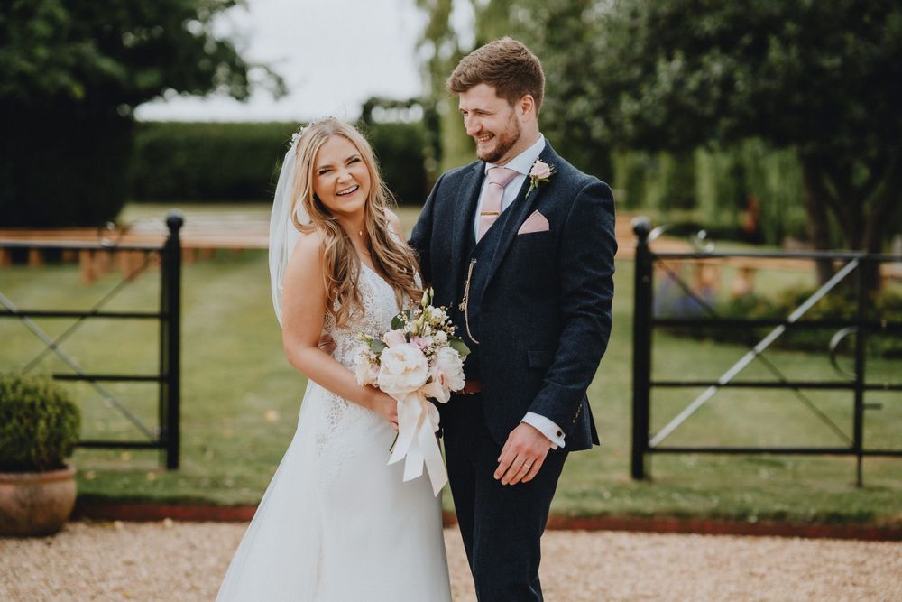 A couple poses together at their outdoor wedding with rustic wooden gates and greenery in the background.