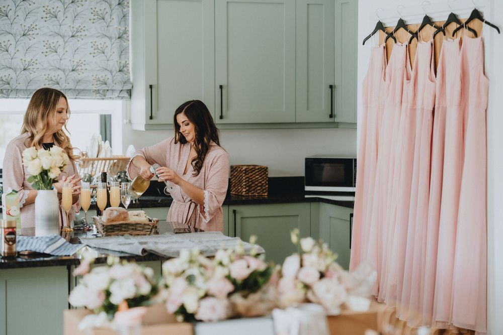 Wedding party members arrange flowers in a kitchen while pink bridesmaid dresses hang nearby.