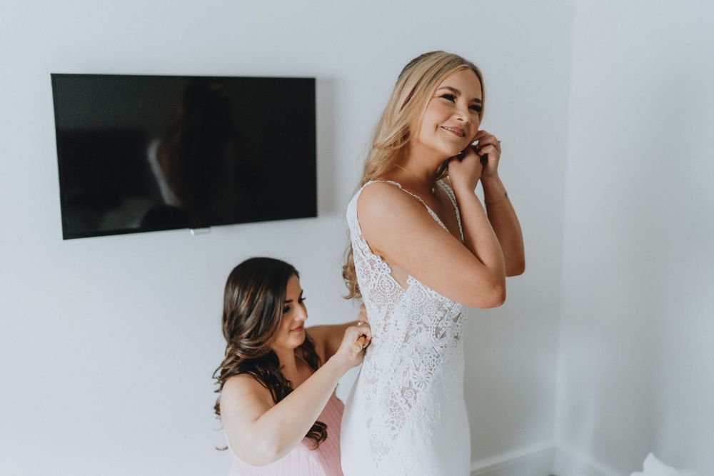 A person helps adjust a beautiful white lace wedding dress during bridal preparations in a bright room.