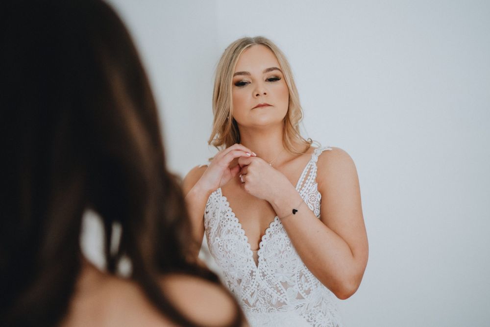 A person in a white lace wedding dress standing in front of a mirror in soft moody lighting.