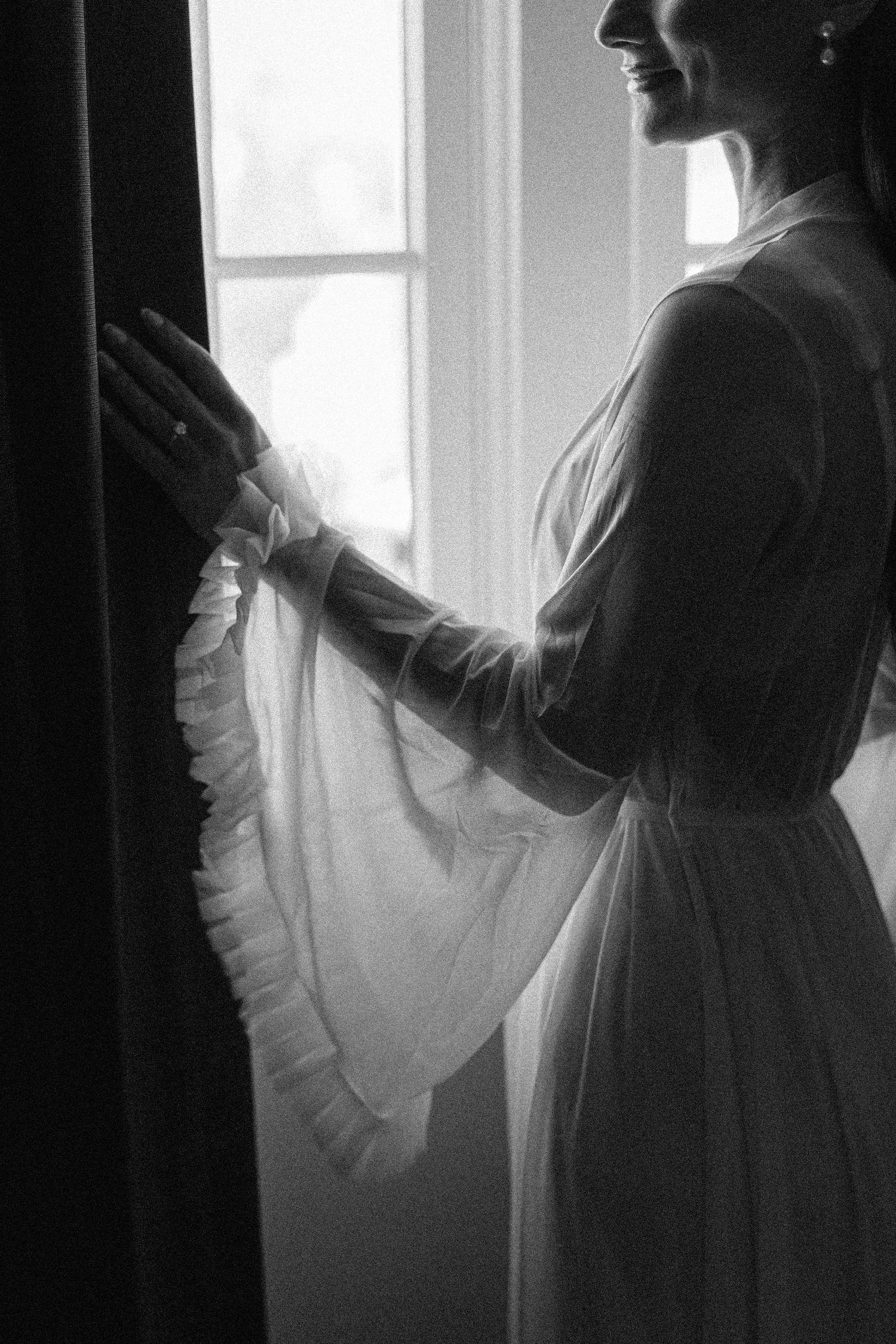 A silhouetted figure in a vintage white dress stands by a window in black and white, creating a delicate, ethereal mood.