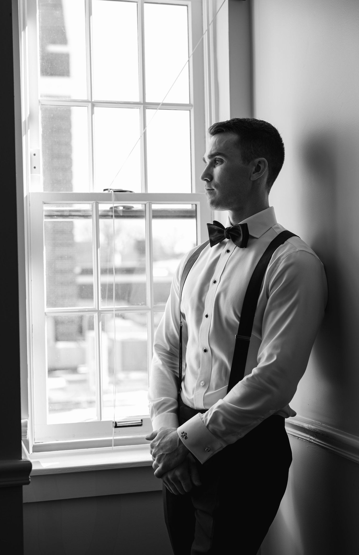 A formally dressed person in suspenders and bow tie stands thoughtfully by a bright window.