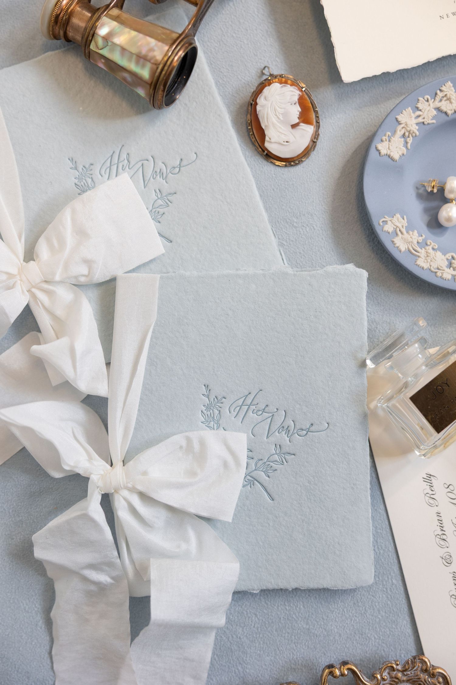 Elegant wedding stationery with silk ribbons, cameo brooch, and vintage decorative elements on a light blue surface.
