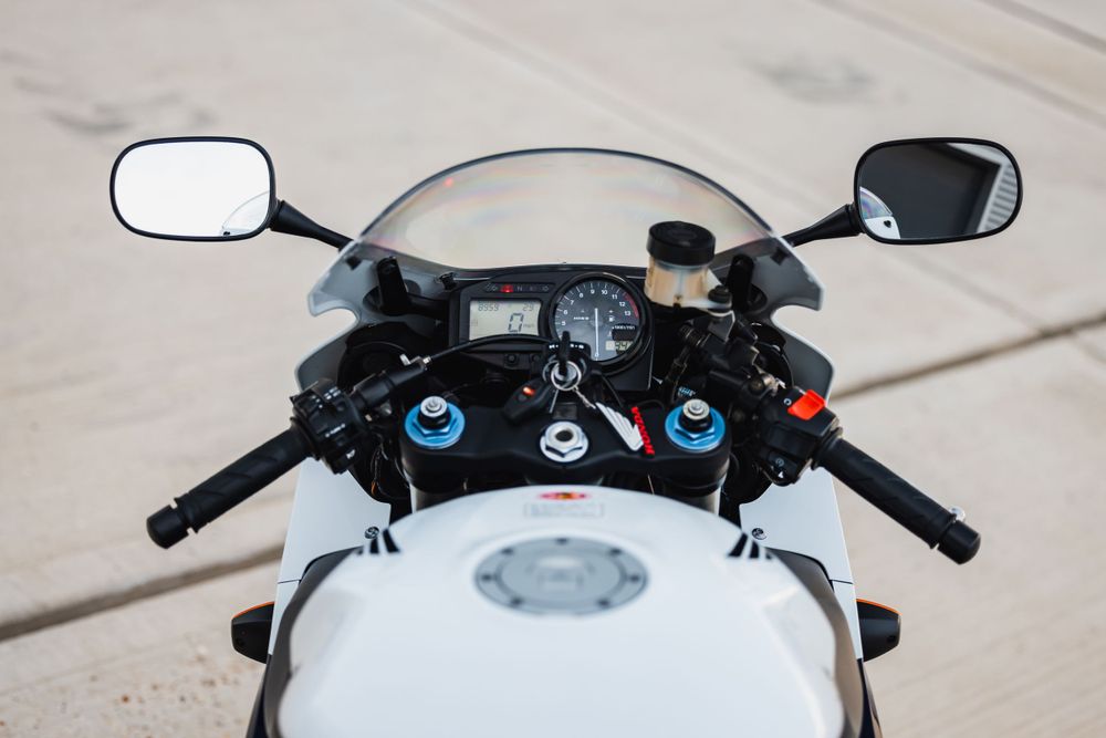Overhead view of white motorcycle handlebars with blue gauges and mirrors mounted on a concrete surface.