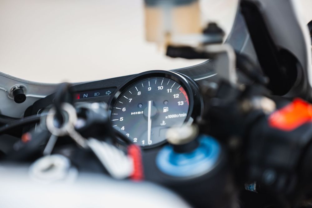 Close-up view of a motorcycle speedometer gauge with blurred background showing partial dashboard details.