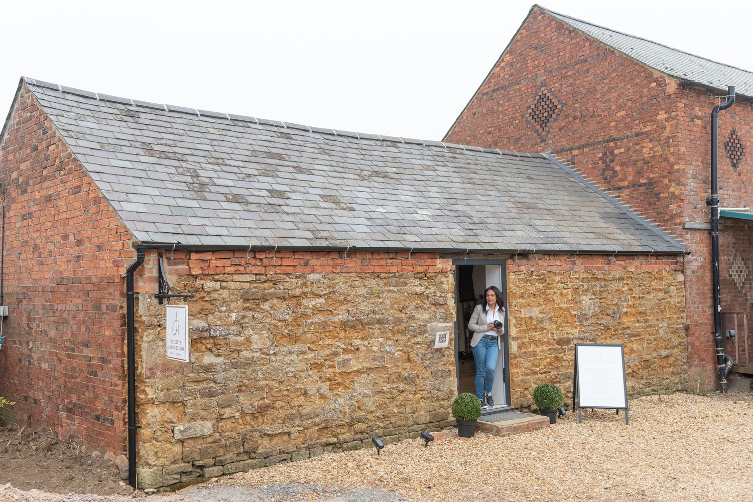 Stone and brick outbuilding with rustic exterior features a person standing in a doorway on a gravel surface.