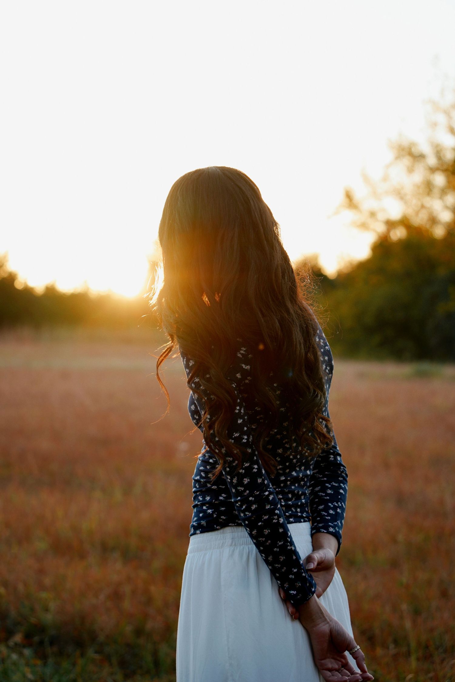Woman in floral top and white skirt holding hands while standing in a golden sunset field.