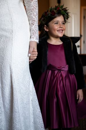 Young flower girl in a burgundy dress and black cardigan stands beside a wedding gown.