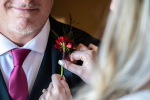 Close-up of someone pinning a red rose boutonniere on a dark suit with purple tie.