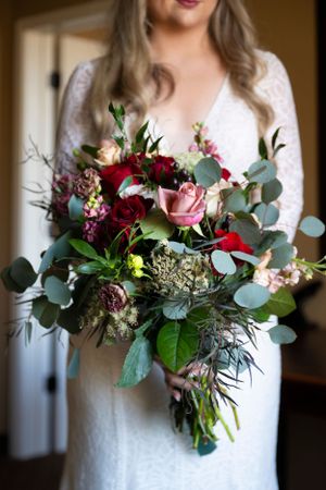 Beautiful wedding bouquet with red roses, pink flowers and eucalyptus held against white lace dress.