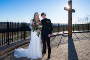 Wedding couple stands near a cross on a scenic overlook with mountains in the background.