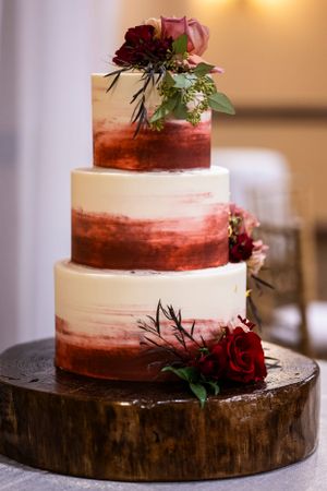 Three-tier wedding cake with burgundy ombre design and red roses on wooden stand.
