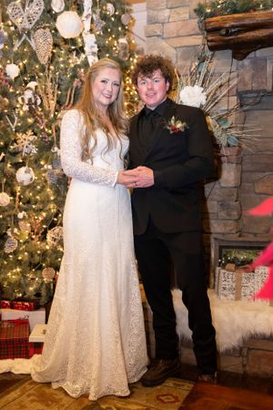 Wedding couple poses in front of decorated Christmas tree and stone fireplace.