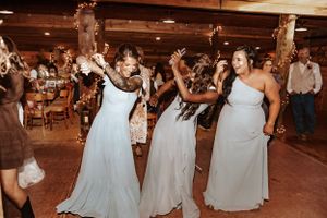 Three bridesmaids in light blue dresses dance together during a wedding reception in a rustic venue.