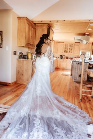 Elegant white wedding dress with long lace train displayed on wooden floor in rustic interior.