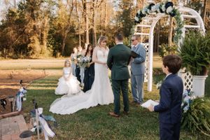 Outdoor wedding ceremony takes place under a white arch decorated with greenery and flowers.