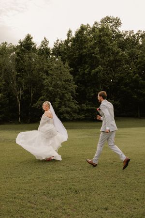 Bride in flowing white gown and groom in light suit running joyfully across green lawn with trees in background.