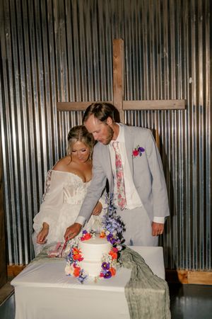 Bride and groom cutting a floral-decorated white wedding cake in front of a rustic corrugated metal wall with a wooden cross.