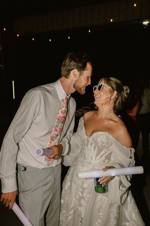 Bride and groom share a romantic moment at wedding reception, holding glow sticks under string lights.