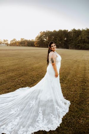 A wedding dress with intricate lace detailing and long train flows across a golden field at sunset.