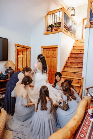 Bridesmaids in pale blue dresses help with wedding preparations inside a rustic wooden staircase area.